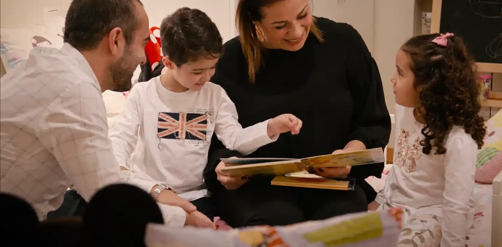 Un padre, una madre y sus dos hijos leen juntos un cuento sentados en la cama, en el vídeo testimonial sobre su proceso con SERMA